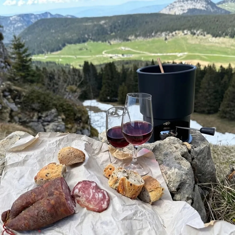 Réchaud à fondue posé sur un rocher face aux montagnes, avec pain, charcuterie et deux verres de vin